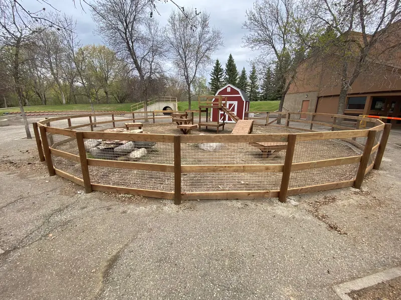 Curved wood rail fence with wire-mesh infill enclosing a fire-pit gathering area near a red barn